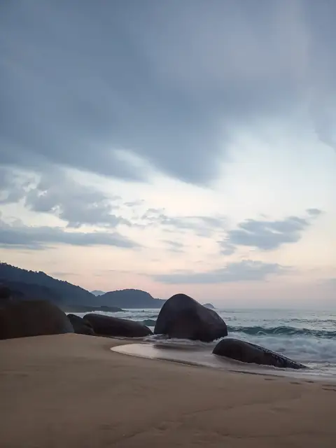 Pedras grandes na areia ao amanhecer, com ondas, morros ao fundo e céu nublado