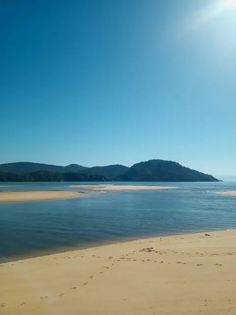 Banco de areia e águas calmas sob céu azul, com morros cobertos de mata ao fundo