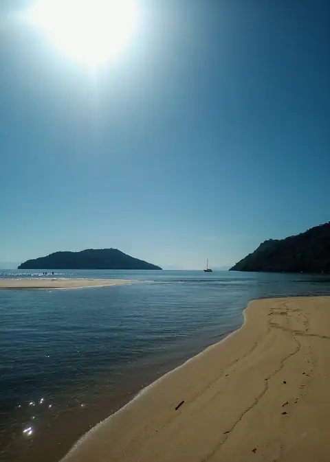 Banco de areia e águas calmas ao sol, com uma ilha e um veleiro ao fundo