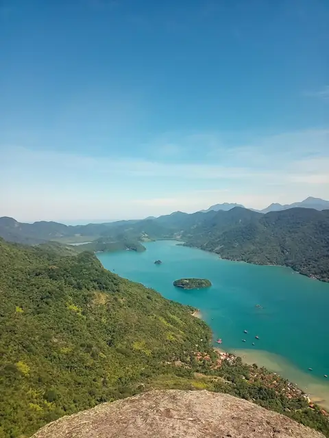 Vista do alto de uma pedra: baía de águas turquesa cercada de mata, com ilhotas, barcos e casas na orla