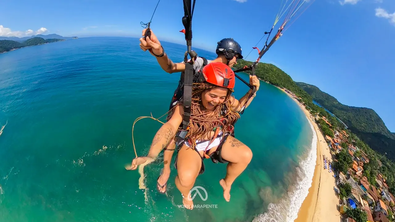 Passageira rindo em voo duplo de parapente sobre uma praia com vila, com o mar verde-azulado ao fundo