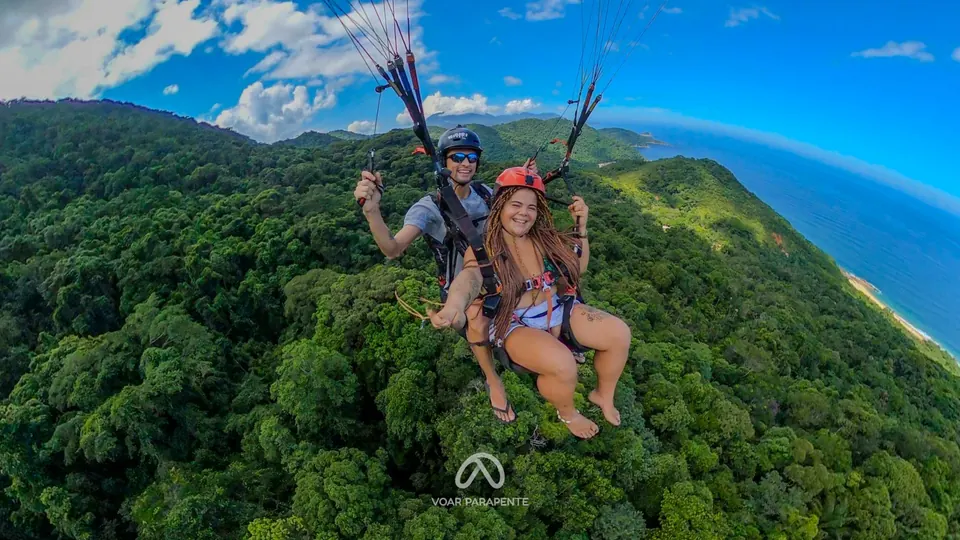 Passageira sorrindo ao lado do piloto, em voo duplo de parapente sobre a Mata Atlântica, com o mar ao fundo