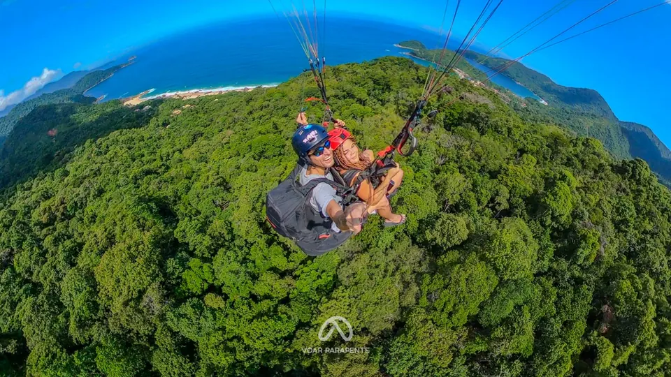 Piloto e passageira em voo duplo de parapente, fazendo selfie sobre a mata, com enseada e morros ao fundo