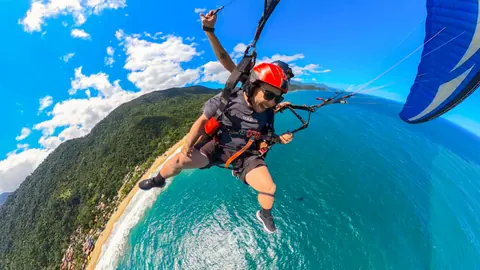Passageiro sorrindo em voo duplo de parapente, com a vela azul ao lado, sobre costão de mata, praia e mar
