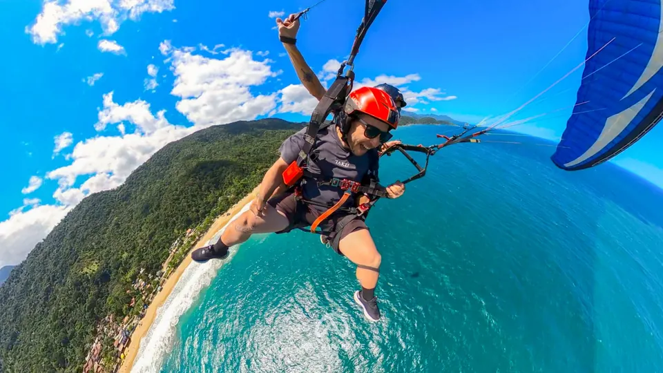 Passageiro sorrindo em voo duplo de parapente, com a vela azul ao lado, sobre costão de mata, praia e mar