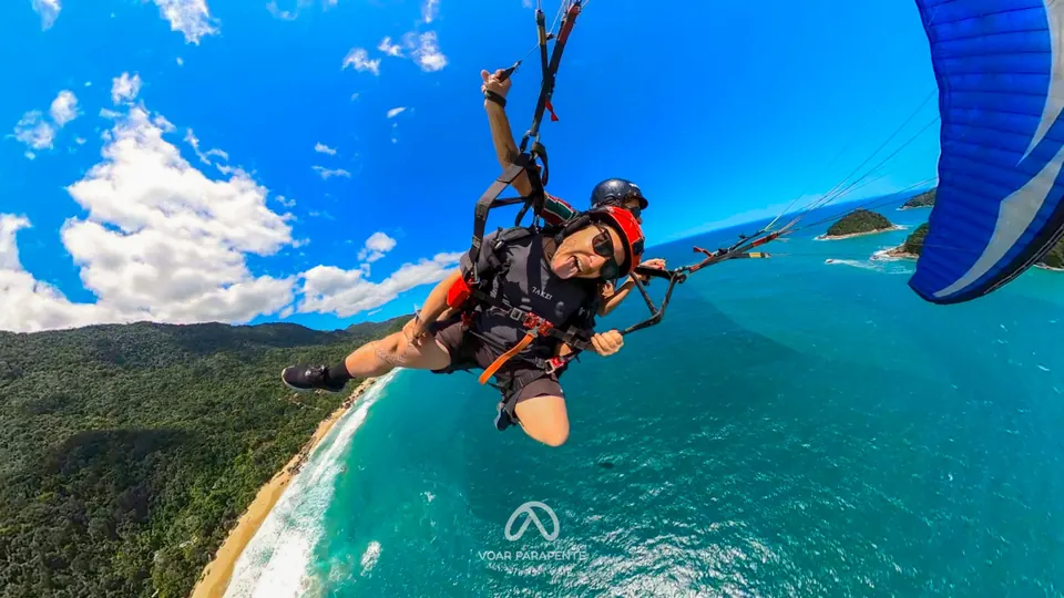 Passageiro de óculos escuros em voo duplo de parapente, com a vela azul ao lado e ilhas ao fundo