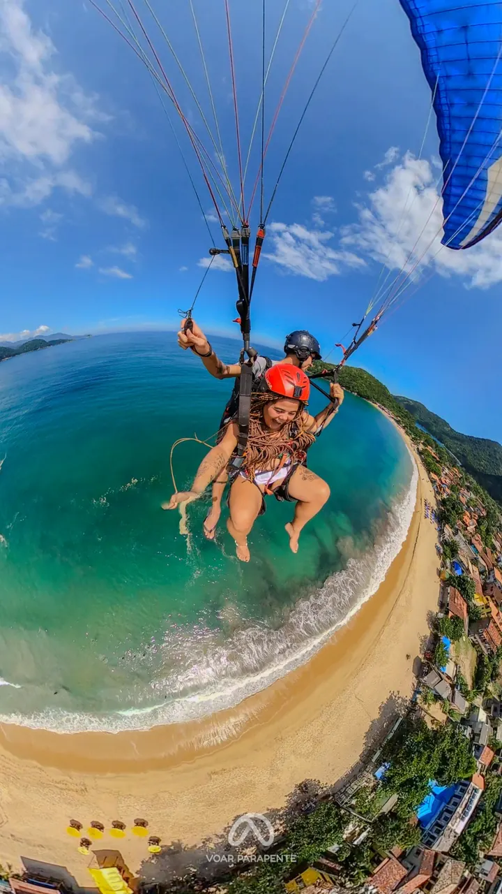Passageira rindo em voo duplo de parapente sobre praia e vila, em imagem vertical com a vela azul no alto