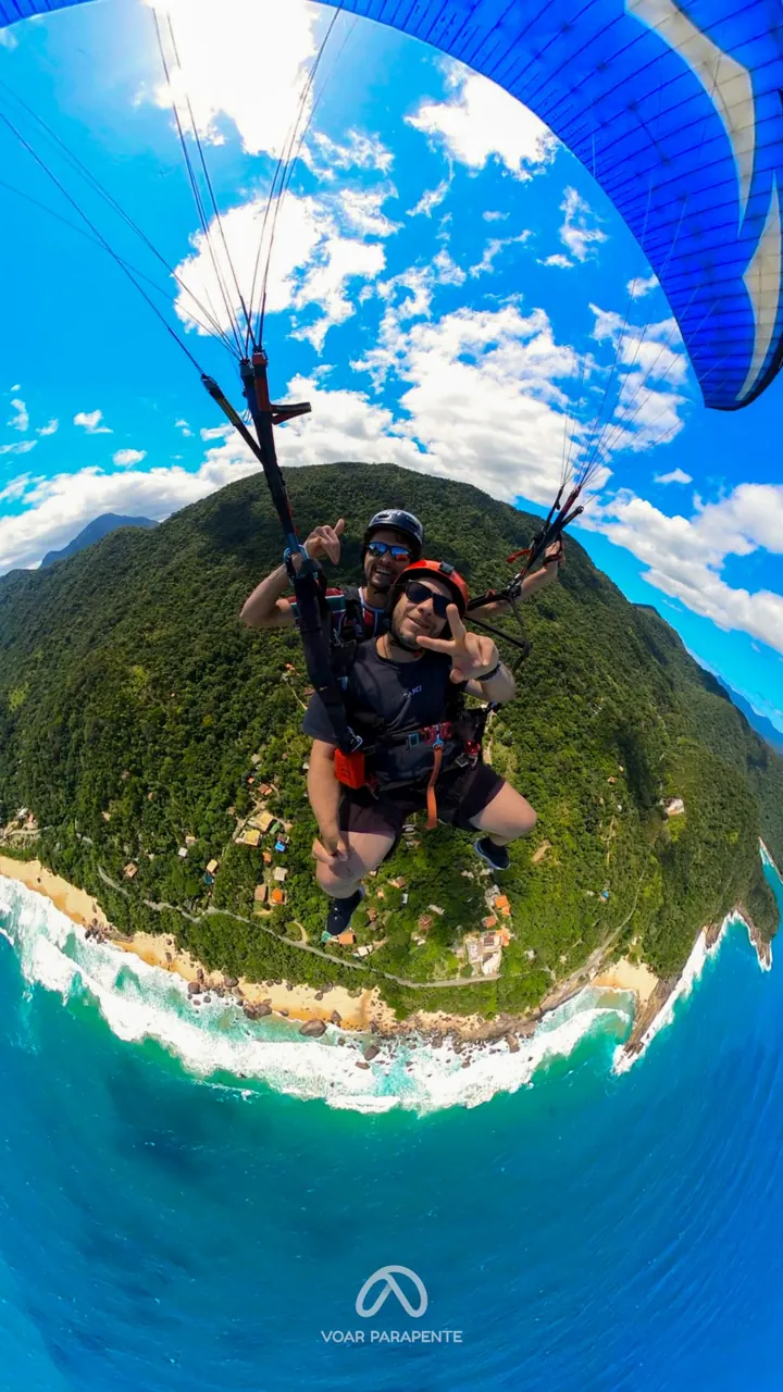 Passageiro fazendo sinal de paz em voo duplo de parapente, sobre uma costa de mata e mar, com a vela azul no alto